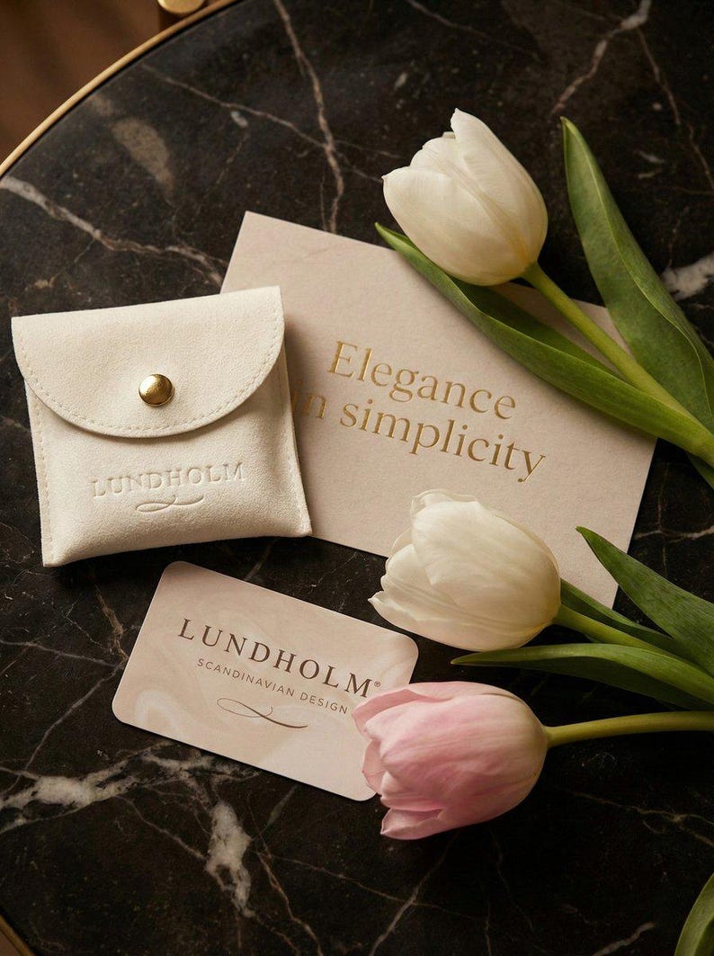 Op de afbeelding: Een cr&egrave;mekleurig su&egrave;de etui met een gouden drukknop, een kaart met de tekst "Elegance in simplicity" en een visitekaartje met de merknaam "Lundholm Scandinavian Design". Witte en roze tulpen zijn gerangschikt op een donker marmeren oppervlak.