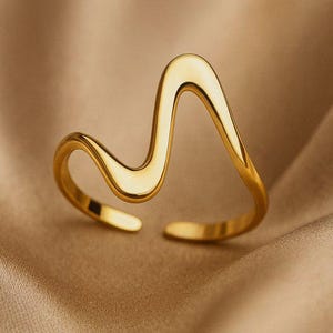 Op de afbeelding: Een goudkleurige, open ring met een golvend ontwerp. De ring heeft een glad, gepolijst oppervlak en een moderne, minimalistische esthetiek. De ring wordt getoond op een neutrale, getextureerde achtergrond.