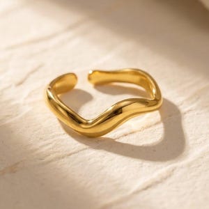 Peut inclure: Une bague ouverte dorée avec un design ondulé. La bague a une surface lisse et polie et est posée sur un fond neutre. Le design de la bague est moderne et minimaliste, adapté au port quotidien.