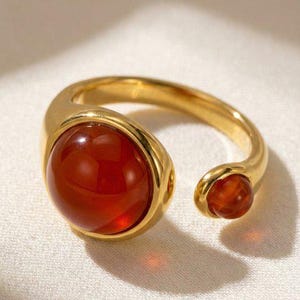 May include: A gold-colored ring with a large, round, reddish-orange gemstone and a smaller matching stone on the open end. The ring has a smooth, polished finish, and the gemstones appear to be cabochon cut.