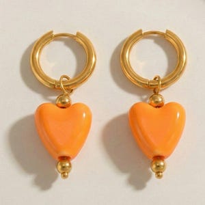 Op de afbeelding: Een paar goudkleurige oorringen met oranje hartvormige hangers. De oorbellen hebben een gepolijste afwerking en een eenvoudig, elegant ontwerp. De hartjes zijn glad en glanzend.