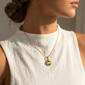 Op de afbeelding: Een goudkleurige ketting met een kleine, ronde hanger met een groene edelsteen. De hanger is gezet in een gouden rand en hangt aan een fijne gouden ketting. De ketting wordt getoond tegen een witte achtergrond.