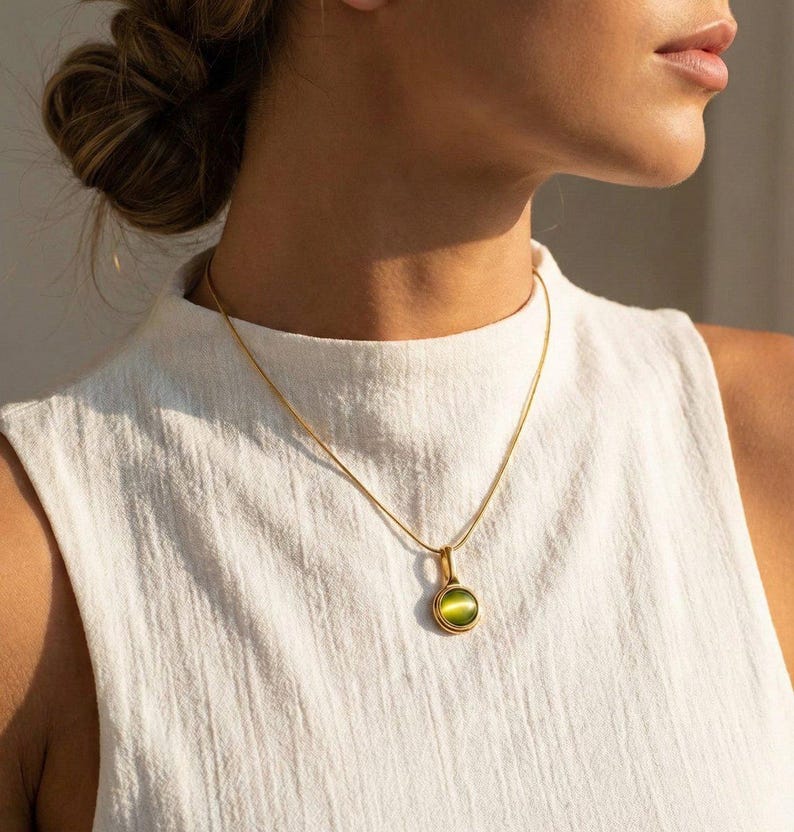 Op de afbeelding: Een goudkleurige ketting met een kleine, ronde hanger met een groene edelsteen. De hanger is gezet in een gouden rand en hangt aan een fijne gouden ketting. De ketting wordt getoond tegen een witte achtergrond.