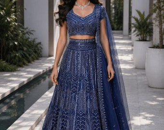 Peut inclure: Un ensemble lehenga choli bleu profond orné de broderies argentées complexes et de sequins sur tout le tissu. La tenue comprend un chemisier à manches courtes, une jupe longue taille haute et un dupatta assorti en voile drapé sur une épaule.