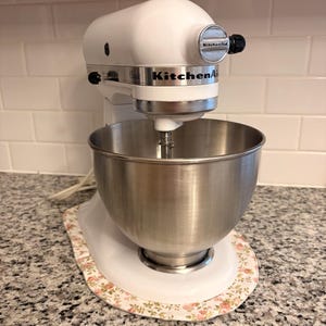May include: A white KitchenAid Classic stand mixer with a stainless steel mixing bowl. The mixer features a silver-toned attachment hub cover with the KitchenAid logo and a black speed control lever on the side of the white metal housing.
