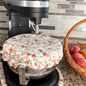 May include: A black KitchenAid stand mixer features a white fabric bowl cover with a floral pattern of small orange and pink flowers and green leaves. The cover has an elasticized edge that fits snugly over the stainless steel mixing bowl.