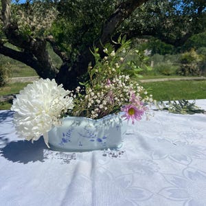 Puede incluir: Un jarrón de cerámica azul claro con estampado floral contiene una variedad de flores, incluyendo un gran crisantemo blanco, pequeñas flores blancas y una margarita rosa. El jarrón está sobre un mantel blanco, con un fondo exterior borroso.