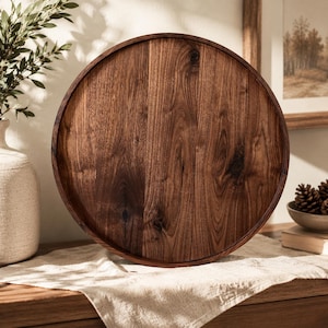 May include: A circular wooden serving tray featuring a rich, dark brown grain pattern with a smooth, polished finish and a raised outer rim. The tray is constructed from solid wood, showcasing natural variations in the wood grain throughout.