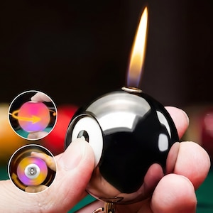 May include: A black and silver novelty lighter shaped like a billiard ball, with a flame at the top. The lighter features a white and black eight ball design. Two inset images show a colour-changing ball and a spinning eight ball.