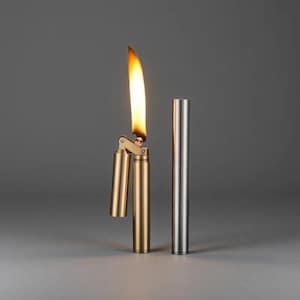 Op de afbeelding: Een messingkleurige aansteker met een open vlam, naast een zilverkleurige cilindrische behuizing. De vlam brandt omhoog. De achtergrond is effen grijs en de aansteker staat op een lichtgrijze ondergrond.