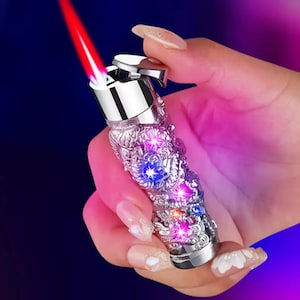 Op de afbeelding: Versierde zilveren aansteker met een rode vlam. De aansteker heeft ingewikkelde draakachtige details en ingebedde kleurrijke lichten. De aansteker wordt vastgehouden in een hand met verzorgde nagels, tegen een donkerblauwe en roze gradiënt achtergrond.