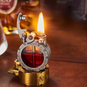Op de afbeelding: Een steampunk-stijl aansteker met een brandende vlam. De aansteker heeft een cirkelvormig, metalen lichaam met een glazen venster dat een rode vloeistof toont. De basis is goudkleurig en de bovenkant is zilverkleurig. De vlam is heldergeel en oranje.