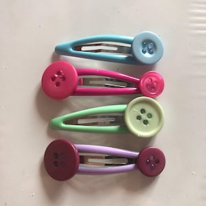 Pode incluir: Quatro grampos de cabelo coloridos com detalhes de botões. Os grampos são em tons de azul, rosa, verde e roxo. Cada grampo tem um botão correspondente, adicionando um toque lúdico aos acessórios de cabelo.