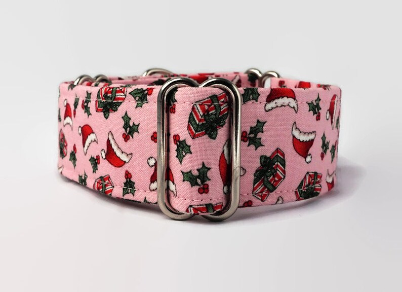 Puede incluir: Collar de perro rosa con estampado navide&ntilde;o con gorros de Pap&aacute; Noel, acebo y regalos. El collar tiene herrajes met&aacute;licos plateados.