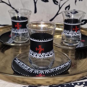 Könnte beinhalten: Klare Teegläser mit schwarzen Bändern und weißen Punkt- und Liniendesigns. Jede Tasse hat ein rotes Symbol. Die Tassen stehen auf einem goldenen Tablett mit passenden Untersetzern.