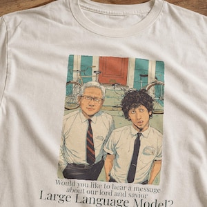以下が含まれることがあります： 白っぽいTシャツ。カラフルなイラストで、白いシャツとネクタイを着た2人の男性が自転車のある建物の前に立っています。Tシャツのテキストは「私たちの主であり救い主であるLarge Language Modelについてメッセージを聞きたいですか？」と書かれています。