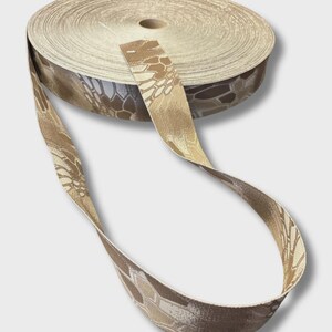 May include: A roll of woven webbing tape featuring a tan, brown, and cream-colored camouflage pattern. The fabric strip is tightly coiled into a circular spool, with one end extending outward to display the repeating hexagonal geometric design.