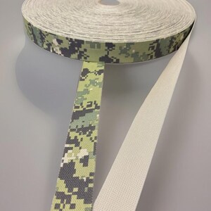 May include: A roll of woven webbing tape featuring a digital camouflage pattern in shades of olive green, dark green, and black on one side, while the reverse side is a solid, plain off-white color with a visible textured weave throughout the material.