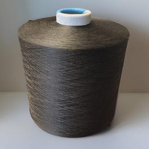 May include: A large cylindrical spool of dark charcoal gray yarn. The spool features a white foam core at the top with a thin blue band visible on the inner rim. The yarn is tightly wound in a cross-hatch pattern around the central tube.
