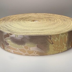May include: A circular spool of heavy-duty webbing tape featuring a multi-colored camouflage pattern in shades of tan, brown, and olive green. The woven fabric has a distinct textured surface and is tightly coiled into a flat, cylindrical shape.