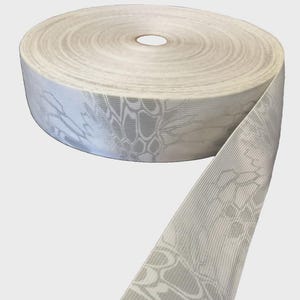 May include: A roll of white woven ribbon featuring a repeating light gray camouflage pattern. The fabric has a textured, ribbed surface with a distinct, interconnected geometric design printed across the entire length of the material.