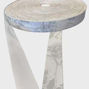 May include: A roll of woven webbing tape featuring a light gray and white hexagonal camouflage pattern. The fabric ribbon has a textured surface with a repeating geometric design, showing a continuous length of material unwinding from the spool.