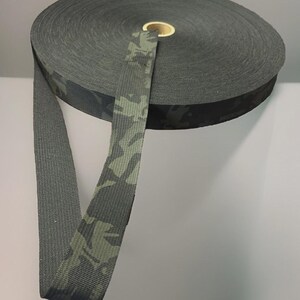 May include: A roll of dark green camouflage webbing. The webbing is a flat, woven material with a repeating pattern of green and black shapes. The roll is cylindrical, with a central cardboard core. The webbing extends from the roll, showing the pattern.