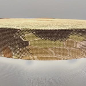 May include: A roll of flat woven webbing strap featuring a multi-colored camouflage pattern in shades of tan, brown, and dark olive green. The material has a tight, textured weave and is coiled into a circular shape with a flat top and bottom.