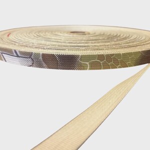 May include: A roll of flat woven nylon webbing tape featuring a camouflage pattern in shades of brown, green, and tan on the outer edge, transitioning to a solid light tan color on the inner coil and the extended length of the strap.