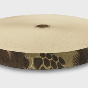 May include: A circular spool of flat webbing tape featuring a tan top surface and a side edge printed with a dark brown and tan camouflage pattern consisting of hexagonal shapes and organic, irregular blotches across the entire outer circumference.