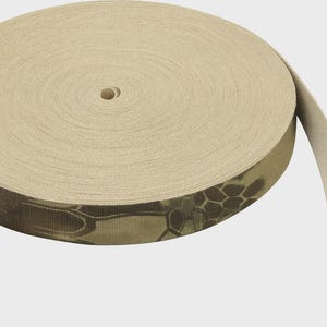 May include: A roll of flat woven webbing tape in a light beige color. The outer edge of the roll features a dark brown and tan hexagonal camouflage pattern. The material has a visible tight weave texture and a consistent width throughout the spool.