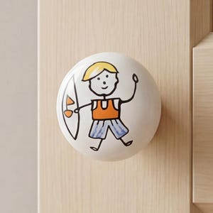 Op de afbeelding: Een ronde witte keramische kastknop met een handgetekende illustratie van een persoon met geel haar, een oranje shirt en een blauwe korte broek, die een wit surfboard met een oranje zeil vasthoudt, gemonteerd op een licht houten kastdeur.