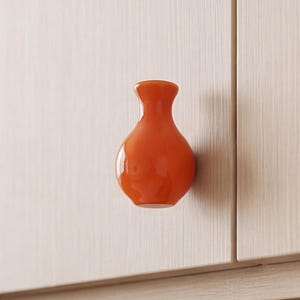 Può includere: Un pomello per mobile di colore arancione brillante e lucido, a forma di piccolo vaso arrotondato con un collo stretto. Il pomello è montato su un'anta di mobile in legno chiaro, creando un contrasto vivace con la superficie neutra e testurizzata.