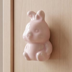 Può includere: Un pomello per mobile in ceramica rosa chiaro a forma di coniglio seduto con orecchie lunghe, testa arrotondata e una finitura liscia e lucida. La figura del coniglio è montata su un'anta di mobile in legno chiaro come maniglia funzionale.