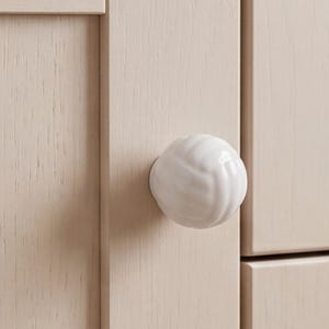 May include: A round, white ceramic cabinet knob with a textured, spiraling knot pattern attached to a light beige, wood-grained cabinet door. The knob features a glossy finish and is centered on the vertical frame of the cabinet door panel.