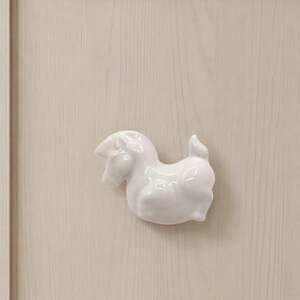 May include: A white ceramic cabinet knob shaped like a stylized horse. The horse figure is depicted in a smooth, glossy finish with a rounded body, a short tail, and a defined head, mounted centrally on a light-colored wooden cabinet door panel.