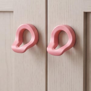 May include: Two light pink cabinet knobs with an irregular, rounded, organic shape. Each knob features a smooth, matte finish and a central opening, mounted onto the light beige, wood-grained doors of a cabinet with a vertical frame design.