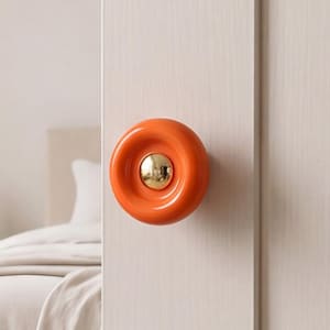 May include: A circular door knob featuring a glossy orange outer ring and a polished gold-tone center dome. The hardware is mounted on a light-colored wooden door panel, showcasing a smooth, reflective finish on both the orange and gold components.