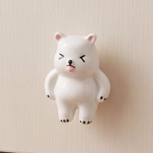Könnte beinhalten: Ein weißer Keramik-Möbelknopf in Form eines stehenden Bären mit verspieltem Gesichtsausdruck. Der Bär hat schwarz bemalte Augen, eine kleine schwarze Nase, eine winzige herausgestreckte rosa Zunge und schwarze Punkte an den Pfoten, montiert an einer hellen Holzschublade.