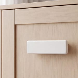 May include: A rectangular white cabinet handle with rounded edges is mounted horizontally on a light-colored wood grain cabinet door. The handle features a smooth, matte finish and is centered on the flat panel of the wooden furniture piece.