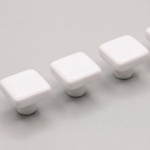 May include: A set of three white square cabinet knobs with a smooth, glossy finish. Each knob features a rounded square top surface and a cylindrical base, arranged in a diagonal line to showcase the reflective, uniform white ceramic material.