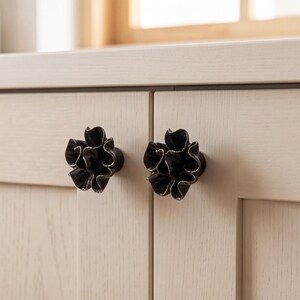 May include: Two dark charcoal gray cabinet knobs shaped like ruffled flowers with layered, wavy petals. These decorative hardware pieces are mounted onto light beige wooden cabinet doors, featuring a glossy finish that reflects light on the edges.