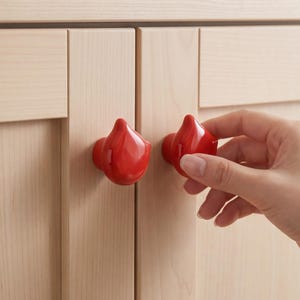 May include: Two glossy, bright red cabinet knobs shaped like stylized teardrops or water droplets. These smooth, rounded knobs are mounted onto light-colored wooden cabinet doors, providing a vibrant contrast against the natural wood grain finish.