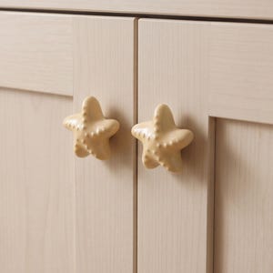 May include: Two light beige, five-pointed starfish-shaped cabinet knobs with a smooth, glossy finish and small, raised textured bumps on the surface. These decorative hardware pieces are mounted onto a light-colored, vertical-grained wooden cabinet door.