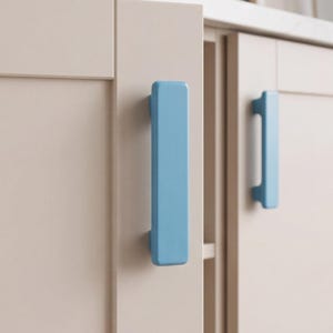 May include: Two rectangular cabinet handles in a matte light blue finish attached to light beige cabinet doors. The handles feature a smooth, elongated design with rounded edges, providing a clean and simple aesthetic for kitchen or furniture cabinetry.