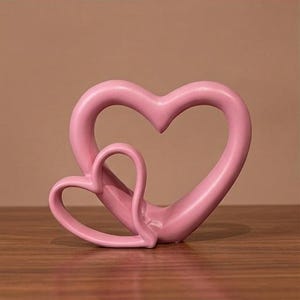 May include: A smooth, matte pink sculpture featuring two interconnected heart shapes. The larger heart frame encompasses a smaller, offset heart, creating a continuous, fluid, and open-work design with rounded edges and a solid, uniform pastel color.
