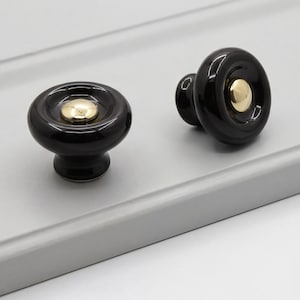May include: Two round cabinet knobs featuring a glossy black finish with a smooth, concave surface. Each knob is centered with a polished gold-tone metal circular inlay, creating a high-contrast look. The knobs have a wide, rounded profile.