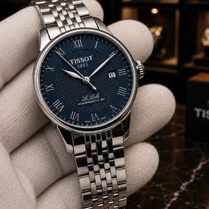 May include: A Tissot Le Locle Powermatic 80 wristwatch with a round silver-tone stainless steel case and a matching multi-link bracelet. The watch features a dark blue textured dial with silver Roman numeral hour markers and a date window.