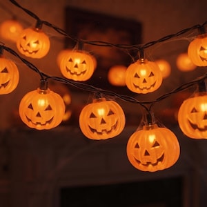 Op de afbeelding: Een lichtsnoer met oranje, pompoenvormige lampjes verbonden door een zwarte draad. Elke pompoen heeft een uitgesneden jack-o'-lantern gezicht met driehoekige ogen en een gekartelde mond. De lampjes geven een warm licht en dunne, kunstmatige spinnenwebben zijn over de draad en de pompoenen gedrapeerd.