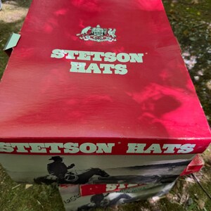 May include: A red Stetson Hats box with white lettering and a logo. The box features an image of a cowboy on a horse. The box is sitting on top of another box with a similar design.